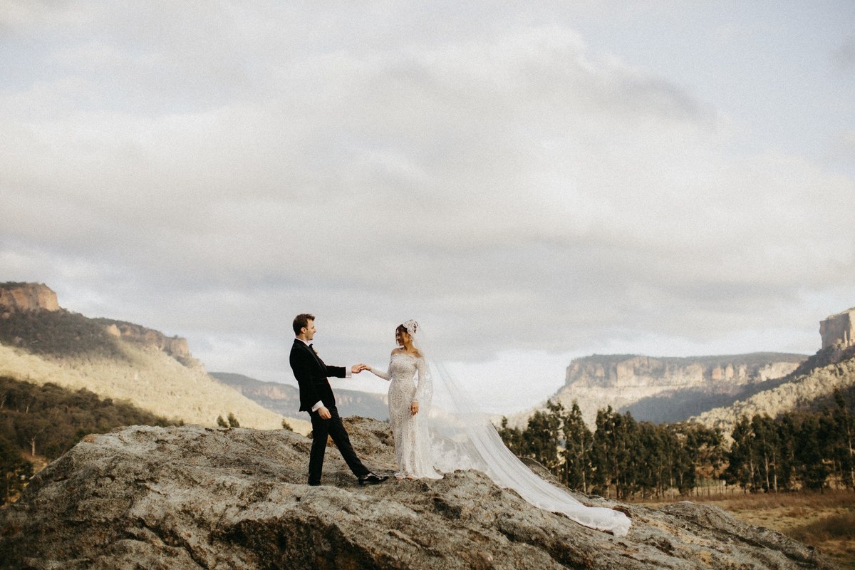 Newlyweds in Wolgan Valley — Joshua Mikhaiel, documentary wedding photographer The bride and groom walk across a rocky ridge above a broad mountain valley. Photograph by documentary wedding photographer Joshua Mikhaiel.