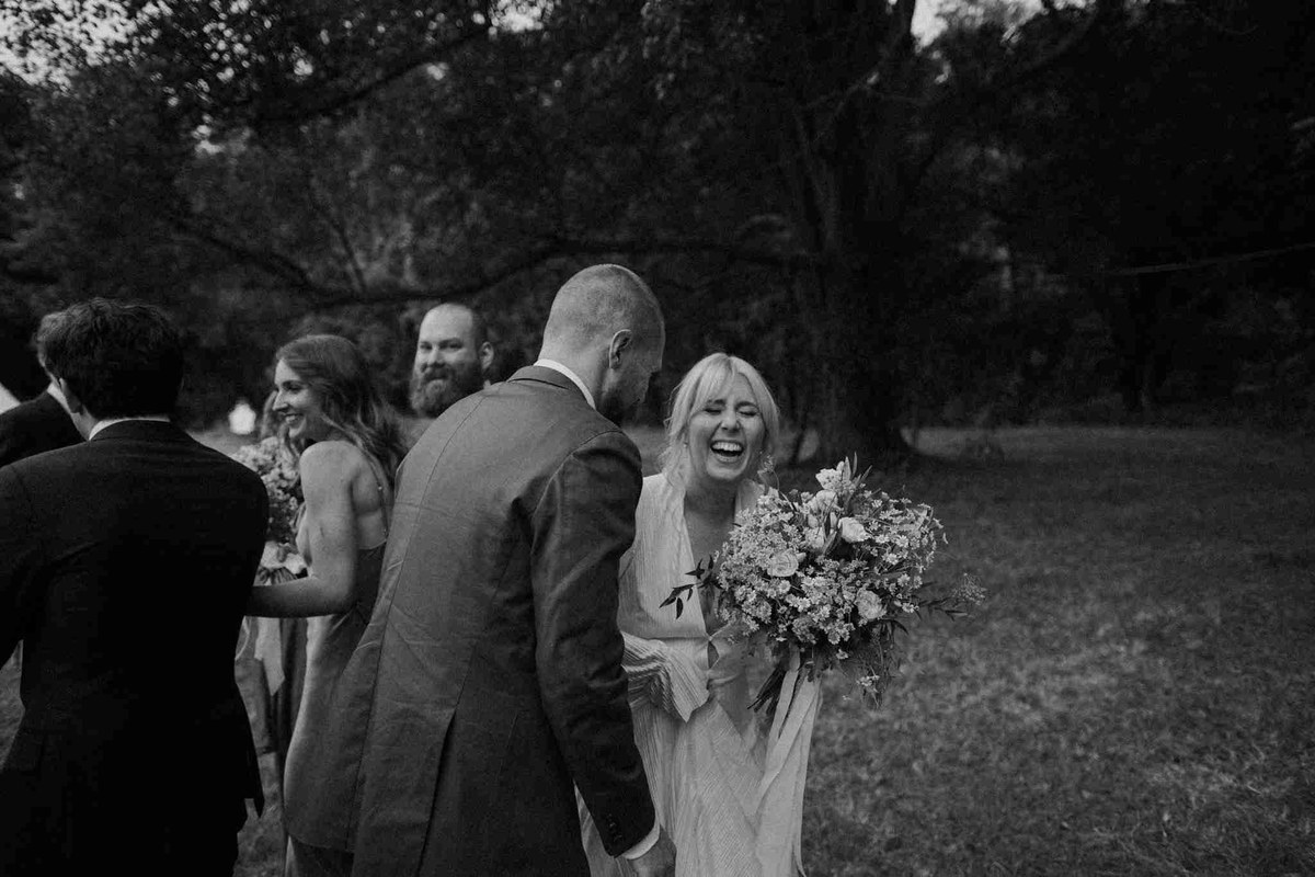 Unposed wedding laughter — Joshua Mikhaiel, documentary wedding photographer A bride laughs with her partner and wedding guests outdoors, in a black-and-white photograph by documentary wedding photographer Joshua Mikhaiel.